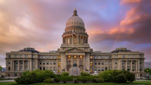 The image shows a grand neoclassical building with a large central dome and two wings, illuminated against a picturesque sunset sky. The structure is surrounded by well-manicured lawns and bushes. The building's facade features tall columns and ornate details, reminiscent of where to find life-changing government jobs in Iowa.