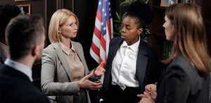 A woman in a beige blouse and gray blazer speaks animatedly to three colleagues in an office with a U.S. flag in the background, perhaps discussing strategies for those wondering how to get promoted in government jobs in Florida. The colleagues, two women and one man, listen attentively, dressed in formal attire. Shelves with books are partially visible.