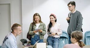 A group of young adults is gathered in a well-lit room. Two people are standing and holding books, one person is sitting at a table with a laptop, and another individual, who appears to be pondering the question "Can Government Jobs in Oregon Help You Pay Off Student Debt?", is standing with a backpack over one shoulder. There are several blue chairs in the background.