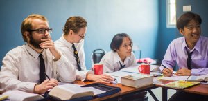 A group of four people sitting at a table, engaged in a study session about career opportunities. They are wearing formal attire with white or light-colored shirts and black ties. Books, notebooks, and red mugs are on the table. The background is a blue wall displaying the question: Can Government Jobs in Oregon Help You Pay Off Student Debt?