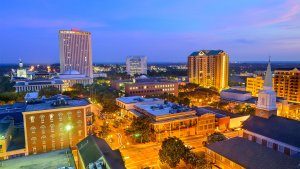A vibrant cityscape at dusk shows a mix of modern and historic buildings, reminiscent of those in Florida. Tall office buildings with lit windows stand amidst shorter structures and tree-lined streets, all illuminated by streetlights. The sky above is painted with shades of blue and purple.