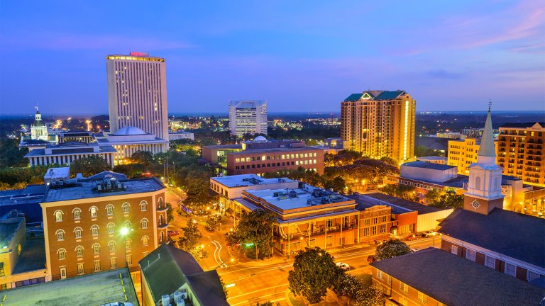 A vibrant cityscape at dusk shows a mix of modern and historic buildings, reminiscent of those in Florida. Tall office buildings with lit windows stand amidst shorter structures and tree-lined streets, all illuminated by streetlights. The sky above is painted with shades of blue and purple.