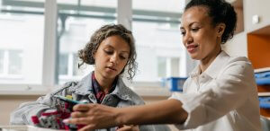 A woman and a young girl work together on a small robotics project at a table. The woman, wearing a white shirt, gestures towards the robot while the girl, in a denim jacket, focuses on the project. They are in a brightly lit room with shelves and a window in the background discussing the best ways to search for government jobs in Colorado.