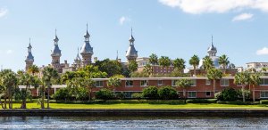 A scenic view of a historic building with multiple silver minaret-style towers, surrounded by lush greenery and palm trees, set against a bright blue sky. Wondering how to get promoted in government jobs in Florida? The foreground features a calm river running alongside modern brick buildings.