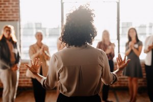 A person with curly hair, seen from behind, gestures with open hands while addressing a small group of people in an industrial-style room with large windows. The diverse group, standing and blurred in the background, appears to be listening attentively or clapping, reflecting a vibrant culture.