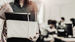 A person holds a box filled with office items, books, and a bouquet of flowers, suggesting their resignation from the workplace. The background features blurred figures of people working at desks in an office setting.