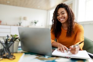 A woman with curly hair and a bright smile sits at a desk in her home office, writing in a notebook while looking at her laptop. She is wearing an orange top. The background, well-lit, features a blurred view of furniture and decorative plants, suggesting she's preparing for her government project.