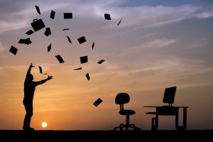 Silhouette of an employee throwing papers and a bag into the air against a sunset sky. A desk, computer, and chair are visible to the right. The image suggests a sense of freedom or release, possibly after completing work or school.