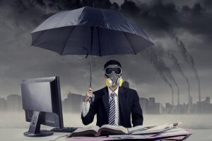 A person in a suit and tie sits at a desk cluttered with papers and an open book, wearing a gas mask and holding an umbrella. A computer monitor, essential for remote work, is on the desk. The sky in the background is dark and smoky with pollution from factory smokestacks emitting smoke.