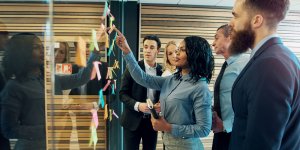 A group of colleagues, both men and women, are gathered around a glass wall covered with sticky notes in an office. One woman is pointing at a sticky note while others are attentively watching, suggesting a meeting or brainstorming session.