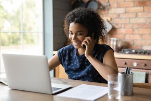 A smiling person with curly hair sits at a wooden table, talking on a smartphone while working on a laptop. Papers, a glass of water, and a pen holder are on the table. Engaged in remote work, they are in front of a modern kitchen with brick walls visible in the background.