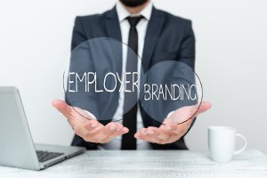 A person in a suit holding two overlapping transparent circles with the words "EMPLOYER" and "BRANDING." Employers' tools, including a laptop and a coffee mug, are on the table in front of them.