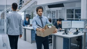 A man in a light blue shirt and tie is carrying a cardboard box filled with office supplies and a small plant, walking through an office. Other employees are seen working at their desks, while one employee is walking away in the opposite direction.