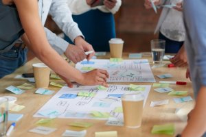 A group of colleagues collaborate on a project at a table. Hands are shown pointing at and arranging papers and sticky notes. Various colored notes and coffee cups are scattered around the wooden surface, indicating a brainstorming session or planning meeting.