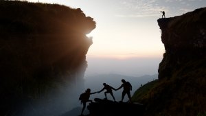 Silhouettes of three people in an action of assisting each other across a rocky gap with one person reaching out from an opposite cliff to help. The sun is setting or rising, casting a warm glow on the scene. In a moment of leadership, another person stands triumphantly on the far cliff.