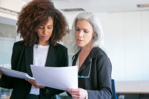 Two women in professional attire, likely employers, are standing in an office closely examining documents. One has curly brown hair and the other has long gray hair. Both appear focused and engaged in their discussion. The background features office furniture and white walls.