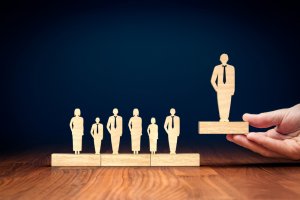 A hand places a wooden figure of a person on a pedestal, elevating it above a row of other wooden figures. All figures are dressed in business attire, representing leadership and distinction in a professional culture. The background is dark, highlighting the figures.