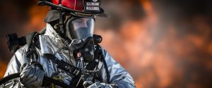 A firefighter in full protective gear, including a red helmet and breathing apparatus, is holding a hose and is focused on fighting a fire with intense orange flames in the background, showing the same dedication you'd need to find high-paying government jobs in Phoenix.