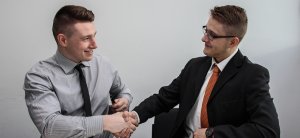 Two businessmen are sitting and shaking hands while smiling at each other. One is wearing a light gray shirt and tie, and the other is in a black suit with an orange tie. They seem to be discussing how to find high-paying government jobs in Phoenix, suggesting a successful strategy meeting or agreement.
