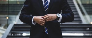 A person in a dark business suit and striped tie stands in front of a staircase, adjusting their jacket. The background includes blurred glass and metal elements, suggesting a modern, professional environment ideal for those seeking high-paying government jobs in Phoenix. The person's face is not visible.