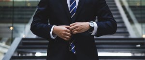 A person in a dark suit and a blue striped tie stands in front of a staircase, adjusting the button on their jacket. The person is wearing a wristwatch and appears to be well-dressed, possibly preparing for an interview on how to find high-paying government jobs in Phoenix.