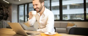 A bearded man in a white shirt is smiling and holding a white coffee cup while sitting at a desk with a laptop. Behind him, there is a modern office with large windows and comfortable seating, perfect for researching how to find high-paying government jobs in Phoenix.