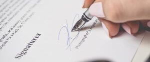 A close-up of a person's hand signing a document with a fountain pen. The document includes the word "Signatures" in bold, and there is space for the photographer's signature. The person's nails, painted light pink, seem to confidently complete this final step after learning how to find high-paying government jobs in Phoenix.