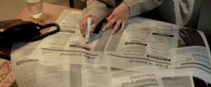 A person wearing a beige sweater is sitting at a table, using a red marker to circle job advertisements in a newspaper titled "How to Find High-Paying Government Jobs in Phoenix." A glass of water and a black telephone with a cord are placed on the table.