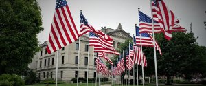 Several American flags are displayed on poles in front of a historic stone building with classical architecture, reminiscent of places where you might discover how to find high-paying government jobs in Phoenix. The building features columns and ornate details, surrounded by trees and greenery, with a pathway leading to the entrance.