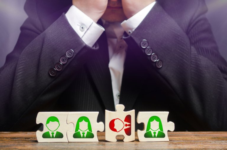 A person in a suit, with their head in their hands, sits behind three puzzle pieces on a table. The first and third puzzle pieces have green icons of people, while the middle puzzle piece has a red icon of a person shouting. This scene captures the challenges of balancing remote work dynamics.