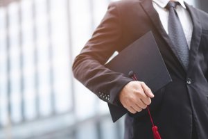 A person dressed in a black suit and tie holds a graduation cap with a red tassel. The background is blurry, featuring tall government buildings. The image conveys a sense of achievement and celebration, likely at a graduation ceremony.