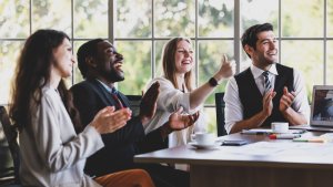 A diverse team of four professionals is sitting at a conference table, smiling and applauding. One woman is giving a thumbs up. The background shows large windows with a view of trees, suggesting a bright and positive atmosphere in a modern office setting.