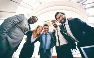 Group of five business professionals dressed in formal attire smiling, standing close together with arms around each other under a bright, modern, curved ceiling. The diverse group appears cheerful and united, reflecting a strong company culture.