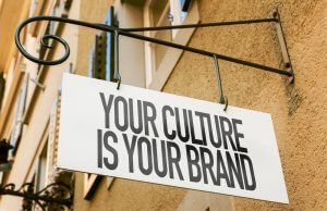 A white rectangular sign with black text that reads "YOUR CULTURE IS YOUR BRAND" hangs from a decorative metal bracket attached to a beige building wall. The background reveals partial views of windows and shutters, subtly highlighting the importance of culture in every detail.