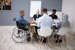 A diverse group of five people in a meeting room sits around a table, engaging in a hybrid meeting. One person is in a wheelchair, and another wears a hijab. Some attendees are using laptops, while others take notes. A whiteboard with charts and post-it notes is visible in the background.