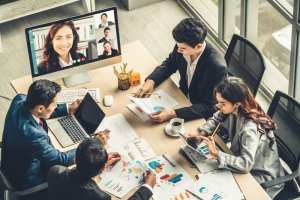 A team of four people in business attire sits around a table covered with charts, graphs, and documents while engaged in a video conference call with a woman displayed on a monitor. The setting is a modern, well-lit office with large windows.