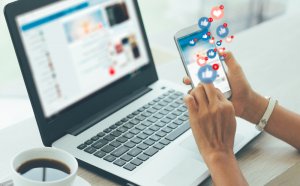 A person uses a smartphone, with social media reaction icons such as likes, hearts, and emojis appearing above the screen. Beside them is a laptop displaying federal news updates and a cup of coffee on the desk, suggesting multitasking between devices.