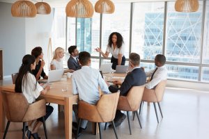 A woman stands and speaks to a group of eight people seated around a table in a modern office with floor-to-ceiling windows. Laptops and papers are on the table, and pendant lights hang from the ceiling, fostering a collaborative culture.
