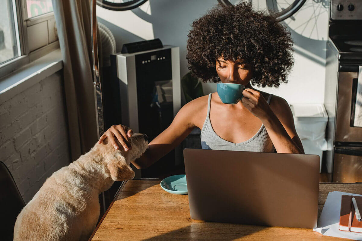 A woman with laptop in her front, drinking from her cup while touching her pet