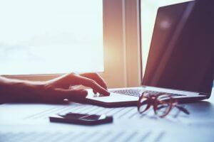 A person using a laptop on a desk near a window. Sunlight streams in, creating a warm atmosphere, as if in a federal office. A pair of glasses and a smartphone are placed on the desk beside the laptop.