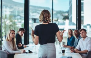A woman stands facing a group of attentive colleagues seated at a conference table in a modern office with large windows. Displaying strong leadership, she appears to be presenting or leading a discussion. The room boasts views of the cityscape and mountains outside.