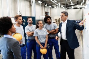 A group of factory workers in gray shirts and blue pants stands in a manufacturing plant, attentively listening to a man in a suit who is demonstrating leadership as he gestures at a whiteboard. One worker holds a yellow hard hat. The environment appears industrial with machinery in the background.