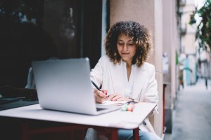 A woman with curly hair sits at an outdoor table working on a laptop. She wears a light blazer and writes something on paper beside the laptop. The setting appears to be a sidewalk café or outdoor workspace, perhaps where professionals discuss governments and policies. The background is blurred.