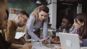 A group of five diverse individuals gathered around a table in a modern office setting. A woman in a blue shirt is standing and pointing to a document on the table, displaying strong leadership skills, while others sit and listen attentively, with a laptop and charts on the table.