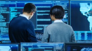Two people are standing in front of large computer screens displaying data and maps, possibly in a control room or data center. They are both looking at a tablet screen, likely discussing the information displayed, alongside their CISO who oversees these critical discussions.
