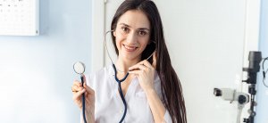 A smiling woman in a white medical coat stands in an office while talking on the phone about high-paying government jobs in Maryland. She holds a stethoscope in her other hand, and medical equipment can be seen in the background.