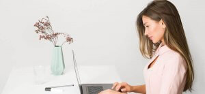 A woman with long brown hair wearing a light pink blouse is sitting at a white table using a laptop, perhaps researching the best resources for finding a federal government job. The table also has a mint green vase with dried flowers, a glass of water, and a pen with a notepad. The background is plain white.