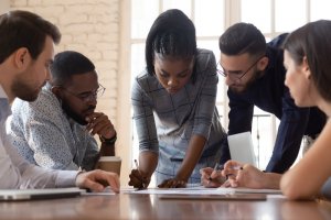A group of five people are gathered around a table, engaged in a discussion. One person in the center is demonstrating leadership by writing on a piece of paper while others attentively look on. The atmosphere appears collaborative and focused.