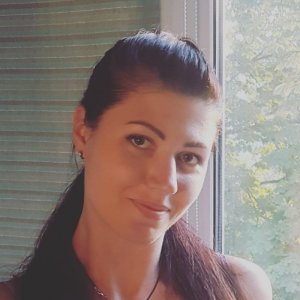 A woman with long, dark hair pulled back smiles softly while standing near a window. Green foliage can be seen outside through the glass. As the CISO of her company, she wears a necklace and appears to be in a well-lit room, exuding confidence and calm.