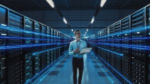 A CISO wearing glasses and a blue shirt holds a tablet while walking through a futuristic data center. Rows of server racks line both sides, with glowing blue lines representing data transfer stretching out in front of and behind the technician.